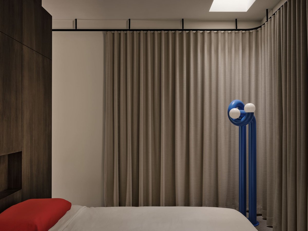 Toronto bedroom with beige curtains, wood paneling, and sculptural blue floor lamp beside a minimalist bed with red pillow