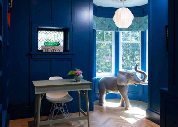 Toronto room with bold blue walls, small desk, bay window seating, elephant sculpture, and natural light creating a playful, modern space
